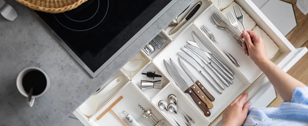Person opening a white kitchen drawer with organized wooden dividers, holding plates, utensils, and ceramic cups
