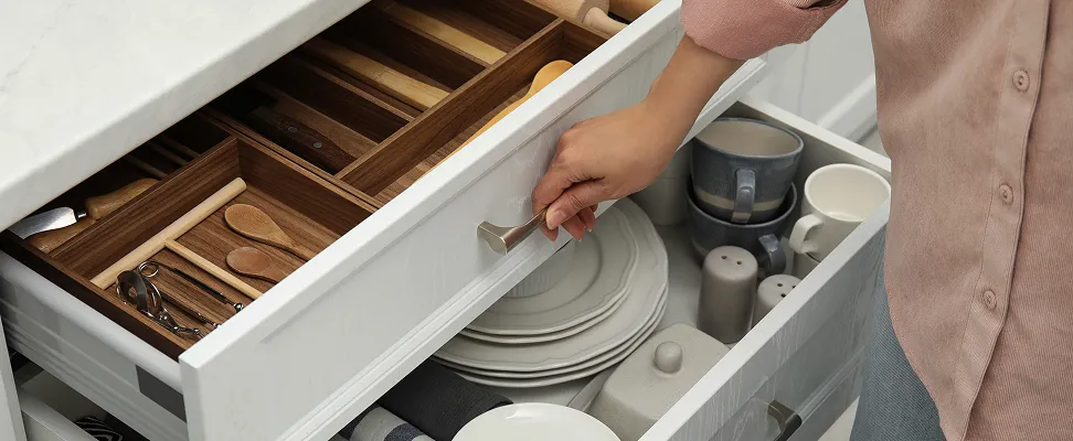Person opening a white kitchen drawer with organized wooden dividers, holding plates, utensils, and ceramic cups